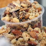 A clear plastic cup filled with a crumbly snack mixture containing almonds, raisins, and chopped nuts, shown on a plate with more of the same in the foreground.